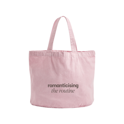 A light pink canvas tote bag with two shoulder straps, featuring the printed text 'romanticising the routine' in dark gray on the front, set against a plain white background.