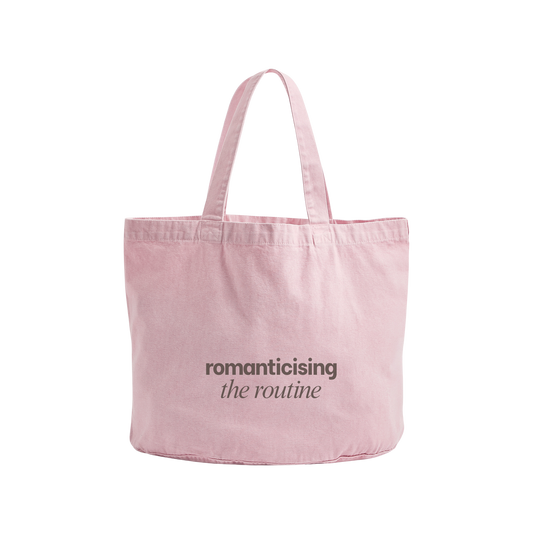 A light pink canvas tote bag with two shoulder straps, featuring the printed text 'romanticising the routine' in dark gray on the front, set against a plain white background.