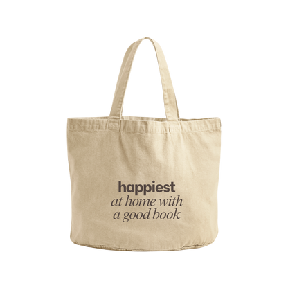 A natural canvas tote bag with the text 'happiest at home with a good book' printed in dark brown on the front, set against a plain white background.