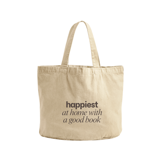 A natural canvas tote bag with the text 'happiest at home with a good book' printed in dark brown on the front, set against a plain white background.