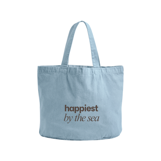 A light blue canvas tote bag with two handles, featuring the text 'happiest by the sea' printed in brown on the front, set against a plain white background.