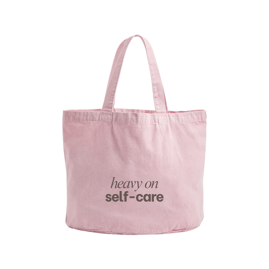 A light pink canvas tote bag with two shoulder straps and the words 'heavy on self-care' printed in dark gray on the front, shown against a plain white background.