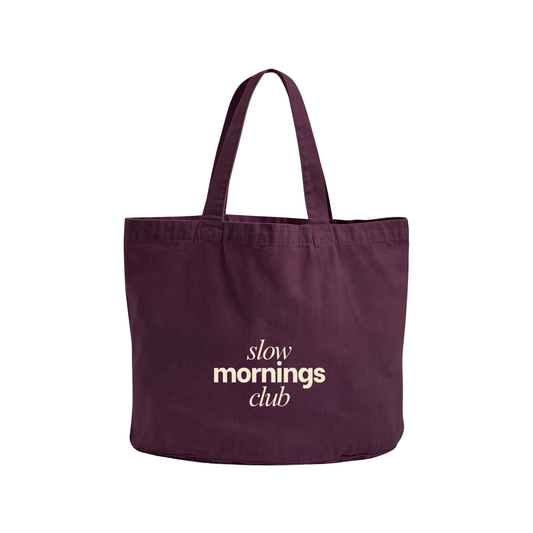 A dark purple canvas tote bag with the words 'slow mornings club' printed in white on the front, set against a plain white background.
